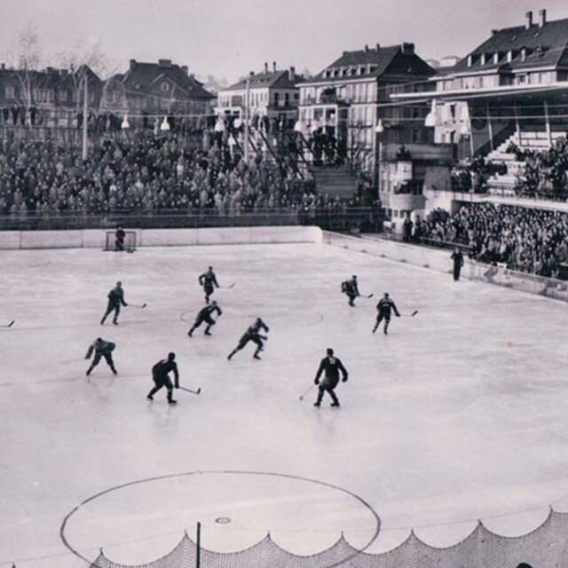 Match de hockey sur glace sur une patinoire extérieure entourée de spectateurs.